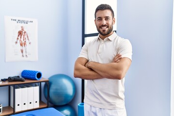 Young hispanic man wearing physiotherapist uniform standing with arms crossed gesture at clinic