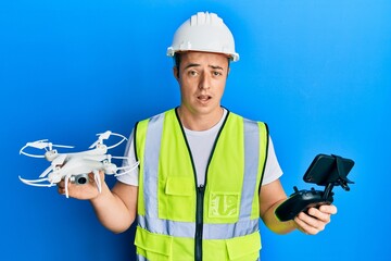 Handsome young man wearing safety hard using drone and control in shock face, looking skeptical and...