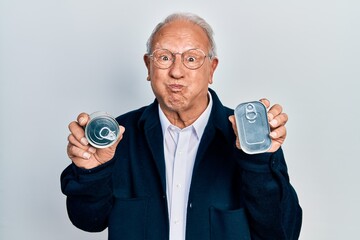 Senior man with grey hair holding canned food puffing cheeks with funny face. mouth inflated with...