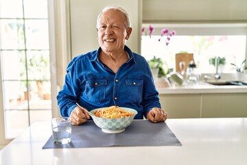 Senior man with grey hair eating pasta spaghetti at home winking looking at the camera with sexy expression, cheerful and happy face.