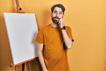 Caucasian man with beard standing by painter easel stand thinking concentrated about doubt with...