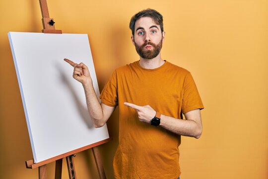 Caucasian man with beard pointing to painter easel stand looking at the camera blowing a kiss being lovely and sexy. love expression.