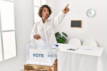 Young hispanic man voting putting envelop in ballot box pointing with finger surprised ahead, open...