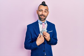 Young hispanic man wearing business suit holding dollar winking looking at the camera with sexy...
