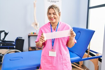 Young hispanic woman working at pain recovery clinic holding bandage winking looking at the camera...