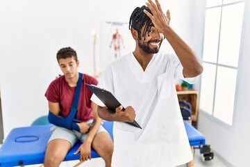Young hispanic man working at pain recovery clinic with a man with broken arm surprised with hand on head for mistake, remember error. forgot, bad memory concept.