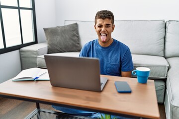 Young handsome hispanic man using laptop sitting on the floor sticking tongue out happy with funny expression. emotion concept.