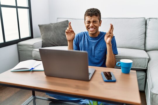 Young handsome hispanic man using laptop sitting on the floor gesturing finger crossed smiling with hope and eyes closed. luck and superstitious concept.