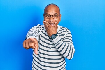 Middle age latin man wearing casual clothes and glasses laughing at you, pointing finger to the camera with hand over mouth, shame expression