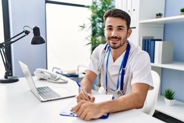 Obraz premium Young hispanic man wearing doctor uniform writing on document working at clinic