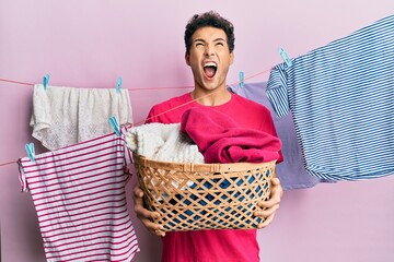 Handsome hispanic man doing laundry holding wicker basket angry and mad screaming frustrated and...