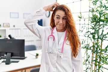 Young redhead woman wearing doctor uniform and stethoscope at the clinic confuse and wonder about question. uncertain with doubt, thinking with hand on head. pensive concept.