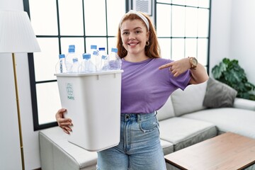 Young redhead woman holding recycling wastebasket with plastic bottles looking confident with smile...