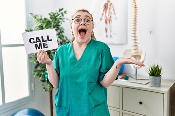 Young redhead doctor woman working at pain recovery clinic holding call me banner celebrating...