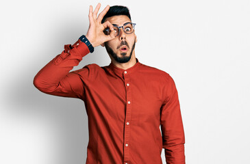 Young hispanic man with beard wearing business shirt and glasses doing ok gesture shocked with surprised face, eye looking through fingers. unbelieving expression.