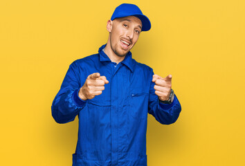 Bald man with beard wearing builder jumpsuit uniform pointing fingers to camera with happy and funny face. good energy and vibes.