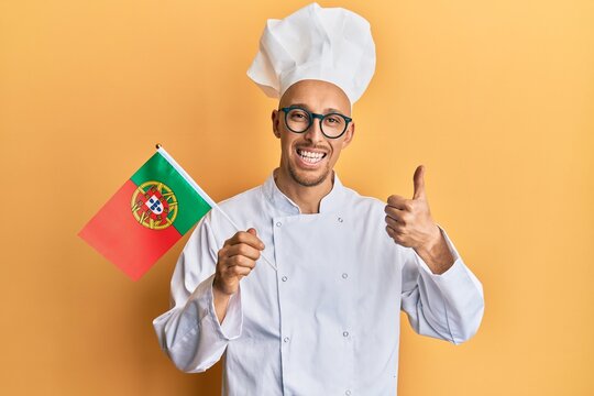 Bald man with beard wearing professional cook apron holding portugal flag smiling happy and positive, thumb up doing excellent and approval sign