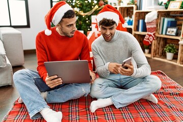 Two hispanic men couple using laptop and smartphone sitting by christmas tree at home