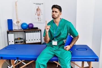 Young physiotherapist man working at pain recovery clinic pointing up looking sad and upset, indicating direction with fingers, unhappy and depressed.