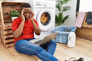 Young african man doing laundry and using computer with hand on head for pain in head because stress. suffering migraine.