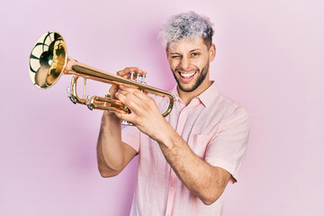Young hispanic man with modern dyed hair playing trumpet winking looking at the camera with sexy...