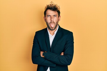 Handsome man with beard wearing business suit with arms crossed gesture and king crown sticking tongue out happy with funny expression.