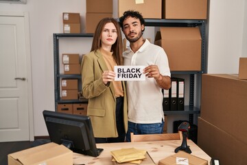 Young two people holding black friday banner at small business store relaxed with serious...