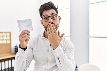 Young man with beard holding covid record card covering mouth with hand, shocked and afraid for...