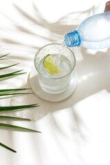 pouring a mineral water from plastic bottle in the glass with lime and ice cubes. Tropical palm leaf shadow. Sunlight and shadow background. beidge minimal.
