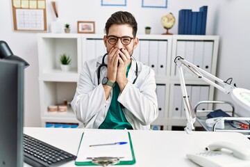 Young man with beard wearing doctor uniform and stethoscope at the clinic laughing and embarrassed giggle covering mouth with hands, gossip and scandal concept