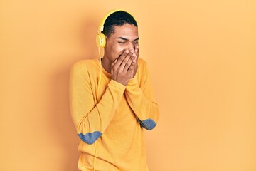 Young african american guy listening to music using headphones laughing and embarrassed giggle covering mouth with hands, gossip and scandal concept
