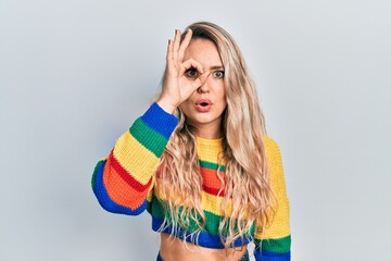 Beautiful young blonde woman wearing colored sweater doing ok gesture shocked with surprised face, eye looking through fingers. unbelieving expression.