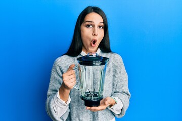 Young hispanic woman holding food processor mixer machine scared and amazed with open mouth for...