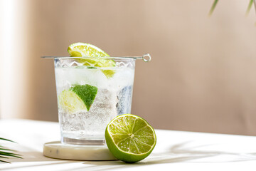glass with cold mineral water with lime and ice cubes. Tropical palm leaf shadow. Sunlight and shadow background. beige minimal.