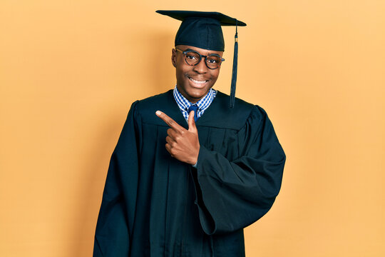 Young african american man wearing graduation cap and ceremony robe cheerful with a smile on face pointing with hand and finger up to the side with happy and natural expression