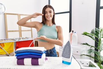 Young hispanic woman ironing clothes at laundry room gesturing with hands showing big and large...
