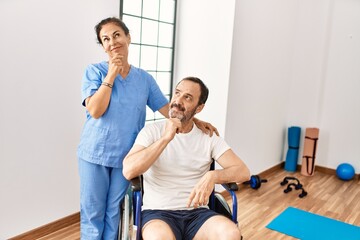 Fototapeta premium Hispanic middle age man sitting on wheelchair and nurse at rehabilitation clinic serious face thinking about question with hand on chin, thoughtful about confusing idea