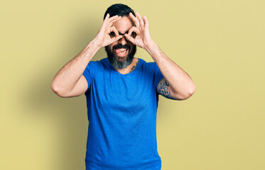 Hispanic man with beard wearing casual t shirt and glasses doing ok gesture like binoculars sticking tongue out, eyes looking through fingers. crazy expression.