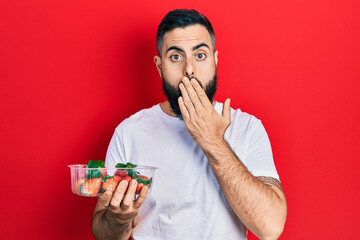 Young hispanic man holding strawberries covering mouth with hand, shocked and afraid for mistake....