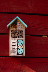 A close up photo of an insect hotel hanging on a red wall outside.