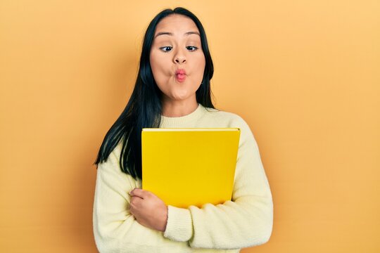 Beautiful hispanic woman with nose piercing holding book making fish face with mouth and squinting eyes, crazy and comical.