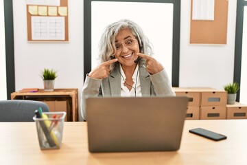 Middle age businesswoman sitting on desk working using laptop at office smiling cheerful showing and pointing with fingers teeth and mouth. dental health concept.