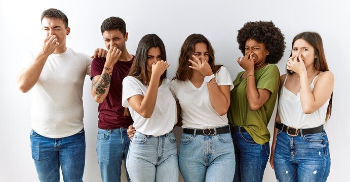 Group of young friends standing together over isolated background smelling something stinky and disgusting, intolerable smell, holding breath with fingers on nose. bad smell