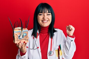 Young hispanic doctor woman holding model of human anatomical skin and hair screaming proud, celebrating victory and success very excited with raised arm