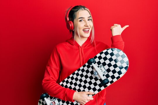 Young modern girl holding skate wearing headphones pointing thumb up to the side smiling happy with open mouth