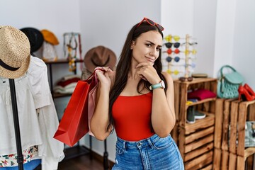 Young brunette woman holding shopping bags at retail shop looking confident at the camera smiling with crossed arms and hand raised on chin. thinking positive.