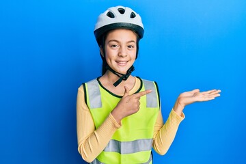 Beautiful brunette little girl wearing bike helmet and reflective vest amazed and smiling to the camera while presenting with hand and pointing with finger.