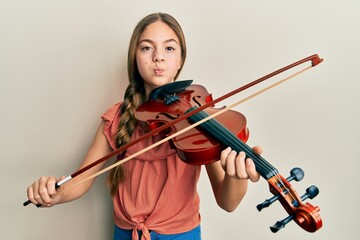 Beautiful brunette little girl playing violin puffing cheeks with funny face. mouth inflated with...