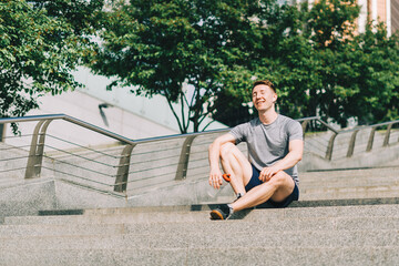 Fototapeta premium Tired Young man runner sitting on stairs and relaxing after sport training. Holding water bottle while doing workout in summer city street