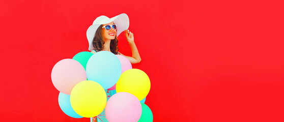 Portrait of happy smiling young woman with colorful balloons wearing summer straw hat on red background, blank copy space for advertising text